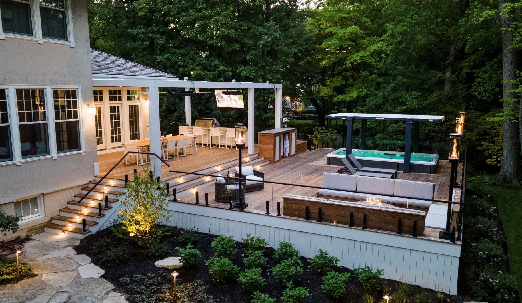 A large deck with a covered portion on the left, a long dining table and several deck chairs. To the right is a hot tub and a seating area with two long benches.