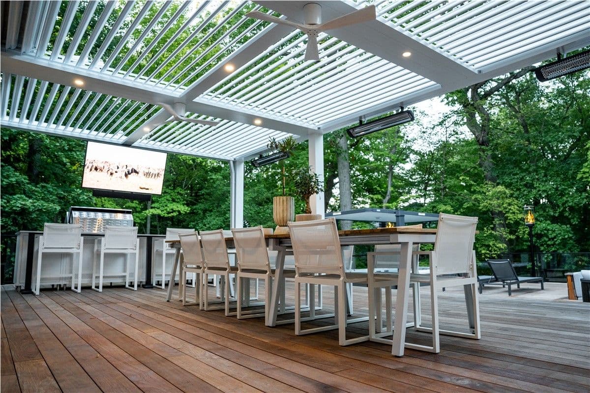 A long table on a deck with white deck chairs. A TV is mounted from crossbeams, and a built-in grill is visible in the background.