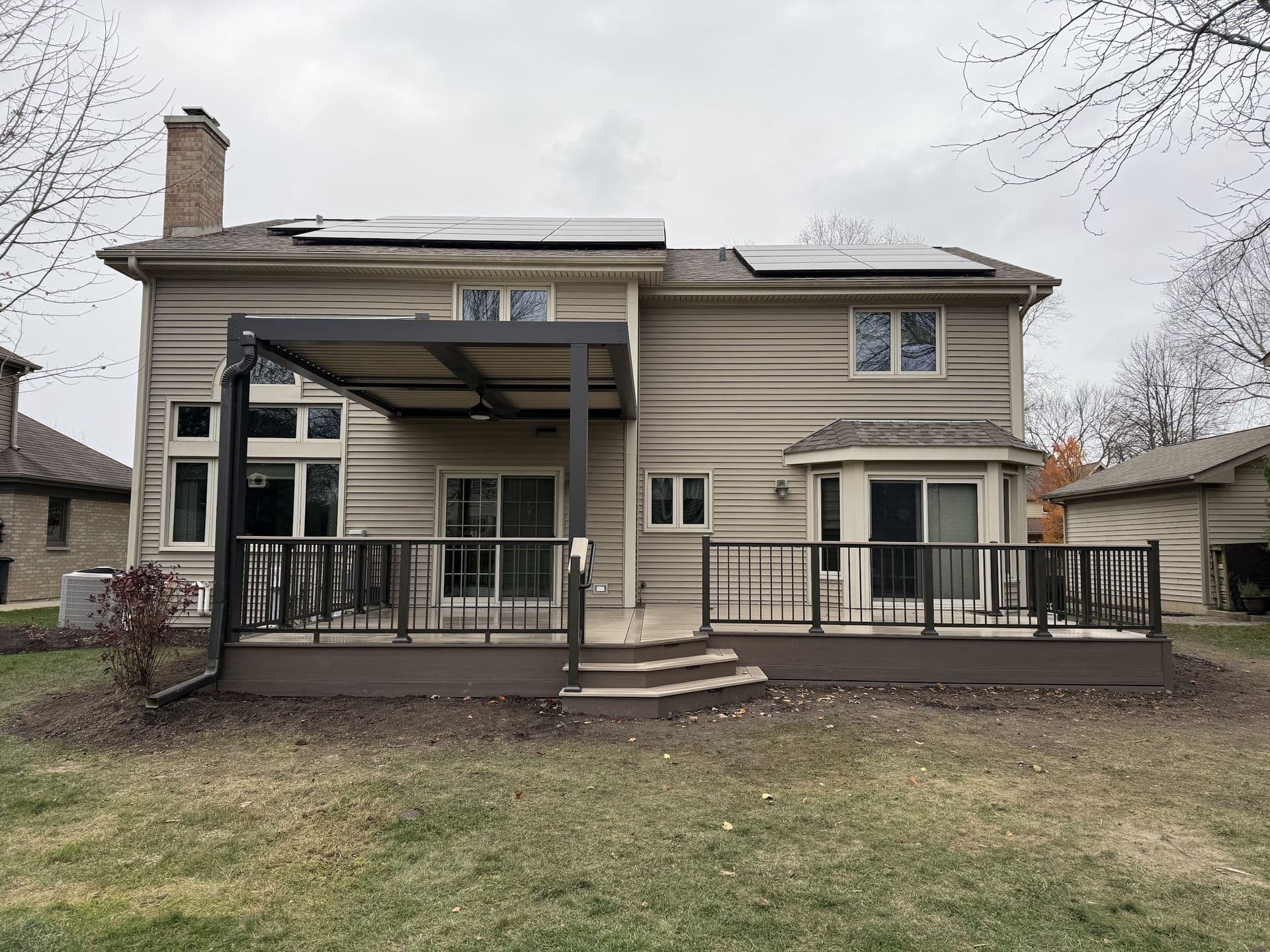 A brown and tan deck off the back of a tan house. The left side is covered with a dark brown pergola.
